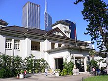 Un bâtiment de deux étages néo-classique montrant influences architecturales japonaises, avec une tour centrale de deux étages. Au premier plan est un court de tennis et jardin et dans le fond des gratte-ciel.