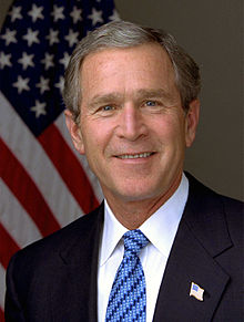 Un coup de portrait d'un homme plus âgé sourire en regardant droit devant. Il a les cheveux courts gris, et porte une veste bleu marine foncé avec une cravate stylé bleu sur une chemise à col blanc. Dans l'arrière-plan est un drapeau américain accroché à un mât.