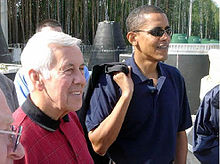 Homme aux cheveux gris et Obama se tiennent, porter des chemises de polo occasionnels. Obama porte des lunettes et détient quelque chose en bandoulière sur son épaule droite.