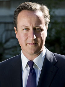 Un homme, rasé de près, avec une courte brun foncé droite balayé les cheveux en arrière vêtu d'une veste de costume, chemise blanche et cravate bleue