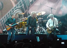 Une photographie en couleur de John Paul Jones, Robert Plant et Jimmy Page sur scène, avec Jason Bonham à la batterie partiellement visible en arrière-plan