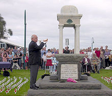 Photo d'un mémorial ANZAC avec un homme âgé à jouer un clairon. Des rangées de personnes sont assises derrière le mémorial. Beaucoup de petites croix blanches avec coquelicots rouges ont été coincé dans la pelouse en rangées de chaque côté du mémorial.