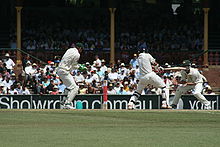 Un match de cricket détenu. Le batteur frappe la balle et les autres joueurs essaient de l'attraper. Le champ vert et le public sont visibles à l'extrême.