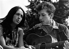 Dylan est assis, en chantant et en jouant de la guitare. Assis à sa droite est une femme regardant vers le haut et chanter avec lui.