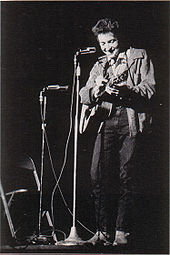 Dylan avec sa guitare sur scène, riant et en regardant vers le bas.