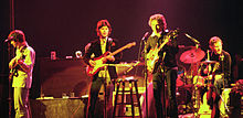 Dylan avec trois musiciens de la bande sur scène. Dylan est le troisième de gauche, vêtu d'une veste noire et un pantalon. Il chante et joue de la guitare électrique.