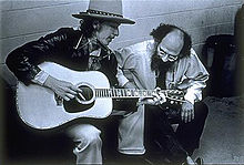 Dylan, vêtu d'un manteau de chapeau et cuir, joue de la guitare et chante, assise. Accroupi à côté de lui est un homme barbu, en l'écoutant, la tête baissée.