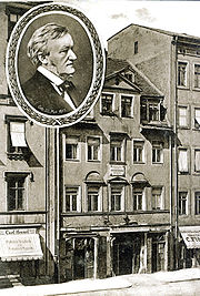 Um cartão de um prédio de cinco andares, com lojas no térreo e janelas sótão no telhado. A inserção rodada tem uma imagem de Wagner na meia-idade.