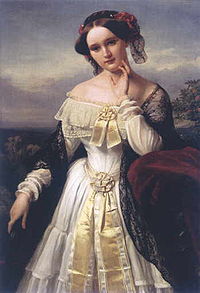 Um retrato do comprimento de três quartos de uma mulher branca jovem ao ar livre. Ela veste um xale sobre um vestido elaborada-de mangas compridas que expõe os ombros e tem um chapéu em cima seu cabelo escuro centralmente parted.
