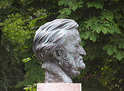 The grey sculpture of a head of a man in his sixties on a plinth with trees in the background. The front of his face is clean shaven but sideburns run under his chin.
