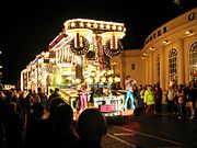 Um grande veÃ­culo iluminado por lÃ¢mpadas multi-coloridas e transportando pessoas em coustumes brilhantemente coloridos na frente de uma grande multidÃ£o em uma rua escura.