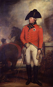Homem barbeado vestindo a jaqueta vermelha de um general do exército britânico 1800 com a estrela da Ordem da Jarreteira, calças brancas, botas pretas de cano alto e um chapéu bicorne preto. Atrás dele, um noivo prende um cavalo.
