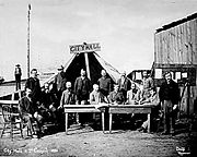 Homens em pé e sentados ao redor de duas tabelas, de frente para a câmera. Uma grande tenda atrás deles tem uma placa de madeira que lê