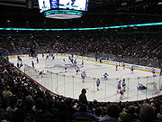 Slightly elevated view of an active ice rink. Players on one team wear mostly red and white uniforms, while the others are outfitted predominately in blue.