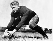 Um jogador de futebol americano uniformizado, mas sem capacete é mostrado em um campo de futebol. Ele está em uma posição de pronto, com as pernas em uma posição larga e ambas as mãos em uma bola de futebol na frente dele.