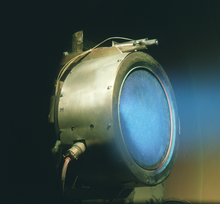 Um cilindro de metal com eléctrodos ligados ao seu lado. Luz difusa azul está vindo para fora do tubo.