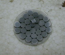 Pílulas cinza escuro em um vidro de relógio. Uma peça cúbica do mesmo material no topo dos comprimidos.