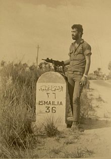 A soldier with an Uzi next to a road sign reading