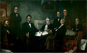 A dark-haired, bearded, middle-aged man holding documents is seated among seven other men.