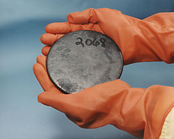 Duas mãos em luvas marrons segurando um disco cinza manchado com um número 2068 escrito à mão sobre ele