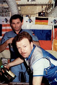 Two men seen in front of a wall featuring a number of switches and dials and covered by large American, Russian and German flags. The men are wearing blue jumpsuits, and two ventilation hoses are visible at the top of the image.