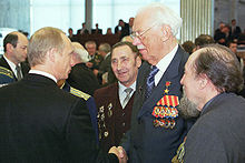 Um homem no centro, de frente para a esquerda, está vestindo medalhas em uma jaqueta. Ele está apertando a mão de um outro homem, observado por outros três.