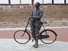 Modern statue of man with moustache in Edwardian cycling clothes holding the handlebar of an ordinary bicycle