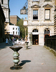 Dois pilares de metal ornamentado com grandes pratos na parte superior em uma rua pavimentada, com uma construção de pedra do século XVIII para trás, sobre a qual pode ser visto as palavras