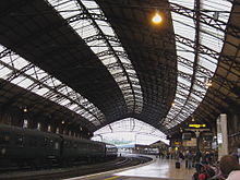A railway station with curved platforms under an arched iron framed roof with roof-lights. A passenger train stands at the platform on the right and on the left passengers waiting for a train.