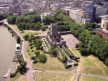 The walls and tower of an old ruined church set in a paved area and surrounded by a park. On the left is water with some pontoons morred and in the background office blocks, streets and church spires.