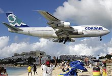 A Corsair 747-300 in white, blue and green livery during landing with its landing gear down, flying over a beach with people directly underneath.