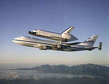 Wide-body airliner in flight with space shuttle mounted on upper fuselage.