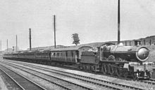 Um retrato preto e branco de quatro linhas ferroviárias em um corte raso, uma grande máquina a vapor leva um comboio de ônibus de meia-esquerda para a direita, em primeiro plano
