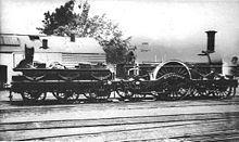 A black and white image showing a steam locomotive facing to the right. The low tender on the left has six wheels; the engine itself has a large wheel in the middle with two wheels in front and one behind.