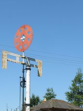A white pole supports two cross-arms joined by four short verticals to make a grid. Above that is a red disc perforated by eight holes.