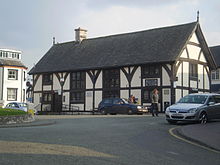 A half timbered building of two floors, with four sets of leaded windows to the front aspect and one set to the side. The build has a steep, slate roof, with a single chimney placed left of centre. Steps and a ramp lead up to its single visible entrance.