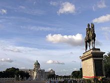 Uma grande praça com uma estátua de bronze de um homem em um cavalo no centro; além da praça é um grande edifício de dois andares com um teto abobadado, janelas e colunas arqueadas