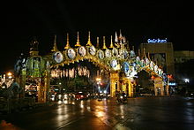An elaborate double archway above a road, with pictures of King Bhumibol Adulyadej; trees decorated with lights