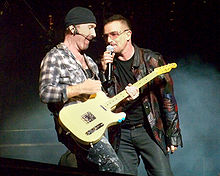 The Edge and Bono stand on a darkened stage, with lit-up smoke behind them. The Edge is strumming a guitar while Bono holds a microphone to his mouth.