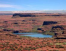 Terreno castanho-avermelhado e muitos pequenos arbustos verdes cercam um lago. Cumes truncadas de rocha escura executado através do terreno paralelo ao horizonte e um ao outro debaixo de um céu azul.