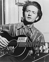 Half-length photo of a middle-aged man sitting in front of a closed door and playing a guitar and singing. His wavy black hair is partly covered by a black hat tipped at a rakish angle. He wears a striped flannel work shirt. His black guitar has a sign on it that says,