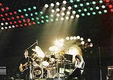 Três membros do grupo durante uma performance ao vivo em Hannover. Da esquerda para a direita, John Deacon (ficou casualmente), Roger Taylor (jogo, sentou-se à bateria), e Brian May (parece estar jogando intensamente). Atrás de Taylor é um tam-tam usado no final de Bohemian Rhapsody. Por trás disso é um grande conjunto de luzes multicoloridas levantadas acima do palco.