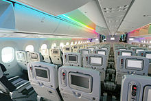 787 cabine. It shows the 787's spacious cabin. Above the blue seats are overhead bins and a rainbow light effect.