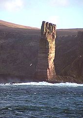 Uma pilha perpendicular altura de rocha marrom está na luz solar na frente de uma costa com altas falésias que se encontram nas sombras.