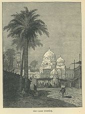 Um homem em um jumento passa por uma palmeira, com uma mesquita e mercado atrás dele.