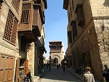 Várias pessoas caminhada por uma pequena pista vazia ofuscado em ambos os lados por edifícios de três andares com varandas e janelas de estilo islâmico envoltas
