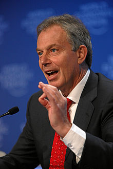 Uma fotografia de um homem com cabelos grisalhos falando em um microfone e gesticulando com a mão esquerda