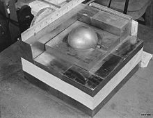 A stack of square metal plates with a side about 10 inches. In the 3-inch hole in the top plate there is a gray metal ball.