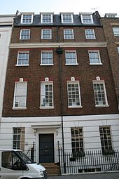 A terrace house with four floors and an attic. It is red brick, with a slate roof, and the ground floor rendered in imitation of stone and painted white. Each upper floor has four sash windows, divided into small panes. The door, with a canopy over it, occupies the place of the second window from the left on the ground floor.