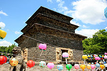 A huge three-story pagoda stands against blue skies. The pagoda is made with bricks of dark gray stone. Colorful lanterns are lined up.